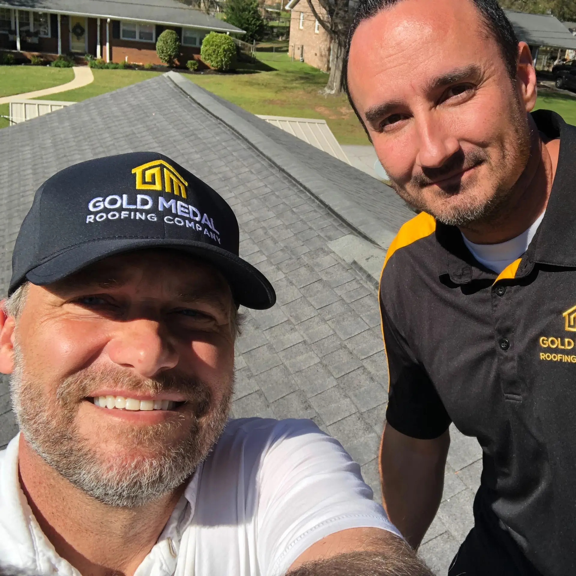 Two roofers from Gold Medal Roofing Company take a triumphant selfie on a recently completed rooftop project in Detroit, Michigan. They proudly wear branded attire, showcasing their team spirit. The man on the left sports a white polo shirt and a black baseball cap with the company's yellow logo, while his colleague wears a black polo shirt with yellow accents. Behind them, a beautifully landscaped house features a sturdy gray roof and a driveway with the company's truck parked, symbolizing their expertise and reliability.
