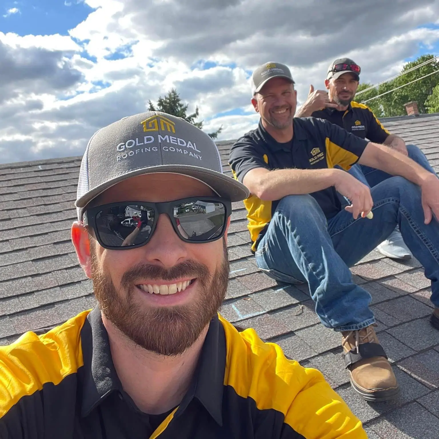 The image features three roofers from Gold Medal Roofing Company, posing on a shingled roof in Erie, Pennsylvania. The foreground roofer, wearing a gray and yellow baseball cap and a black and yellow shirt, takes a selfie with a smile. Behind him, two colleagues sit on the roof, one giving a thumbs up and the other holding a chalk, both wearing matching attire. The background showcases a serene blue sky with white clouds, with trees and power lines visible in the distance.