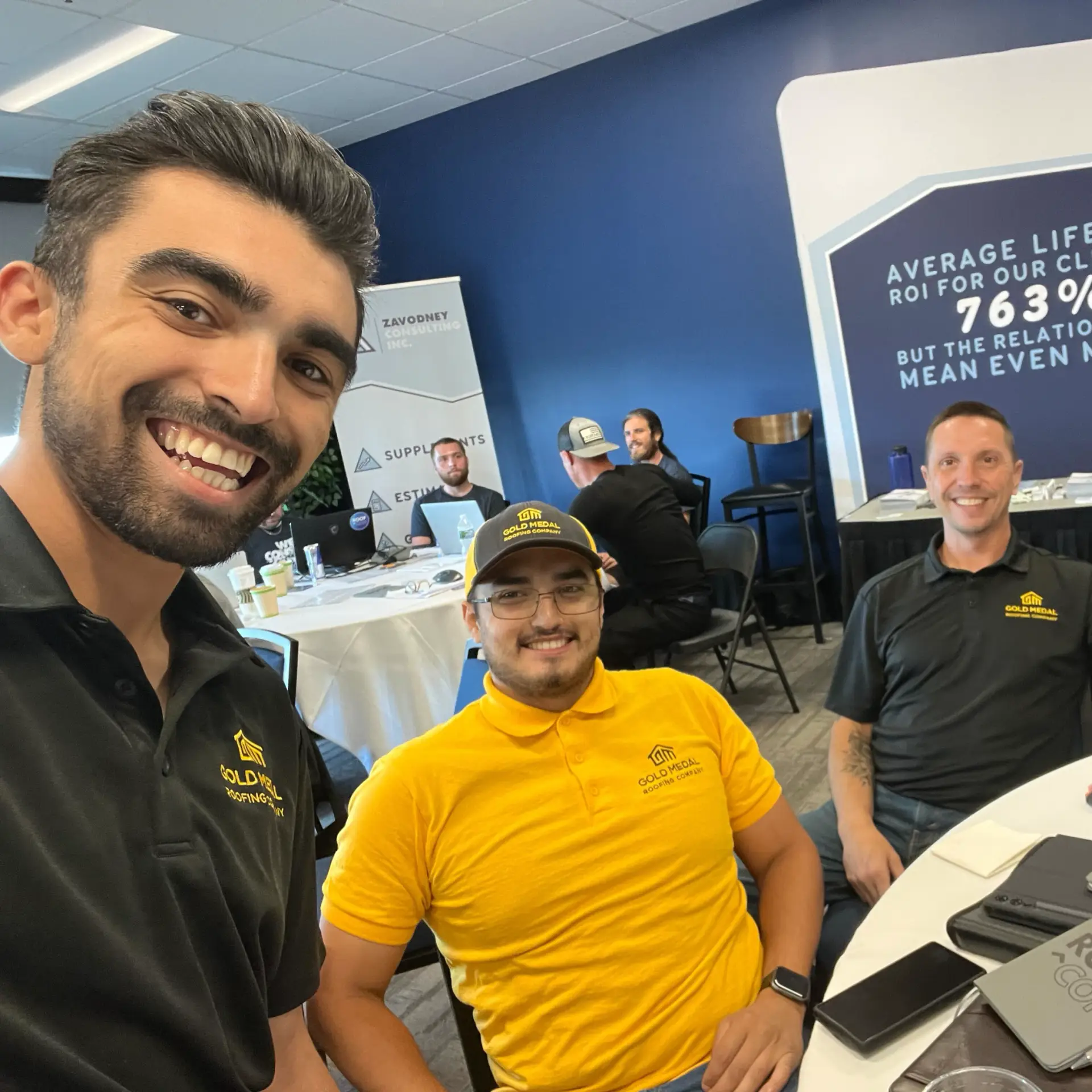 In this image, three roofers, are seated at a table in a conference room, surrounded by a professional setting. The man on the left wears a black polo shirt with a yellow logo, while the man in the middle sports a yellow polo shirt with a black logo and a matching baseball cap. The man on the right is dressed in a black polo shirt with a yellow logo. The table is cluttered with various items, including a drink, a cell phone, and a pamphlet titled "ROOF CON." In the background, a blue wall features a prominent sign that reads, "AVERAGE LIFETIME ROI FOR OUR CLIENTS: 763% BUT THE RELATIONSHIPS MEAN EVEN MORE!"