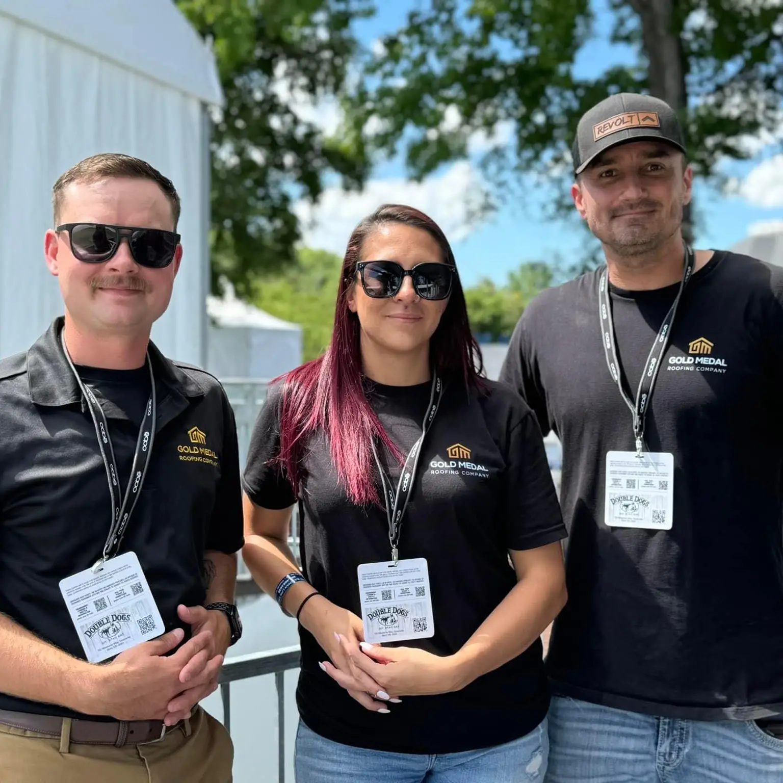 Gold Medal Roofing Company's team poses together in Greenville, South Carolina. Three smiling roofers stand united, proudly wearing branded attire. The man on the left sports a black polo shirt, while his colleagues wear matching black t-shirts with the distinctive "GOLD MEDAL" logo. Khaki pants, blue jeans, and lanyards complete their casual yet professional looks. A crisp blue sky with wispy clouds and a white tent surrounded by trees provides a serene backdrop.