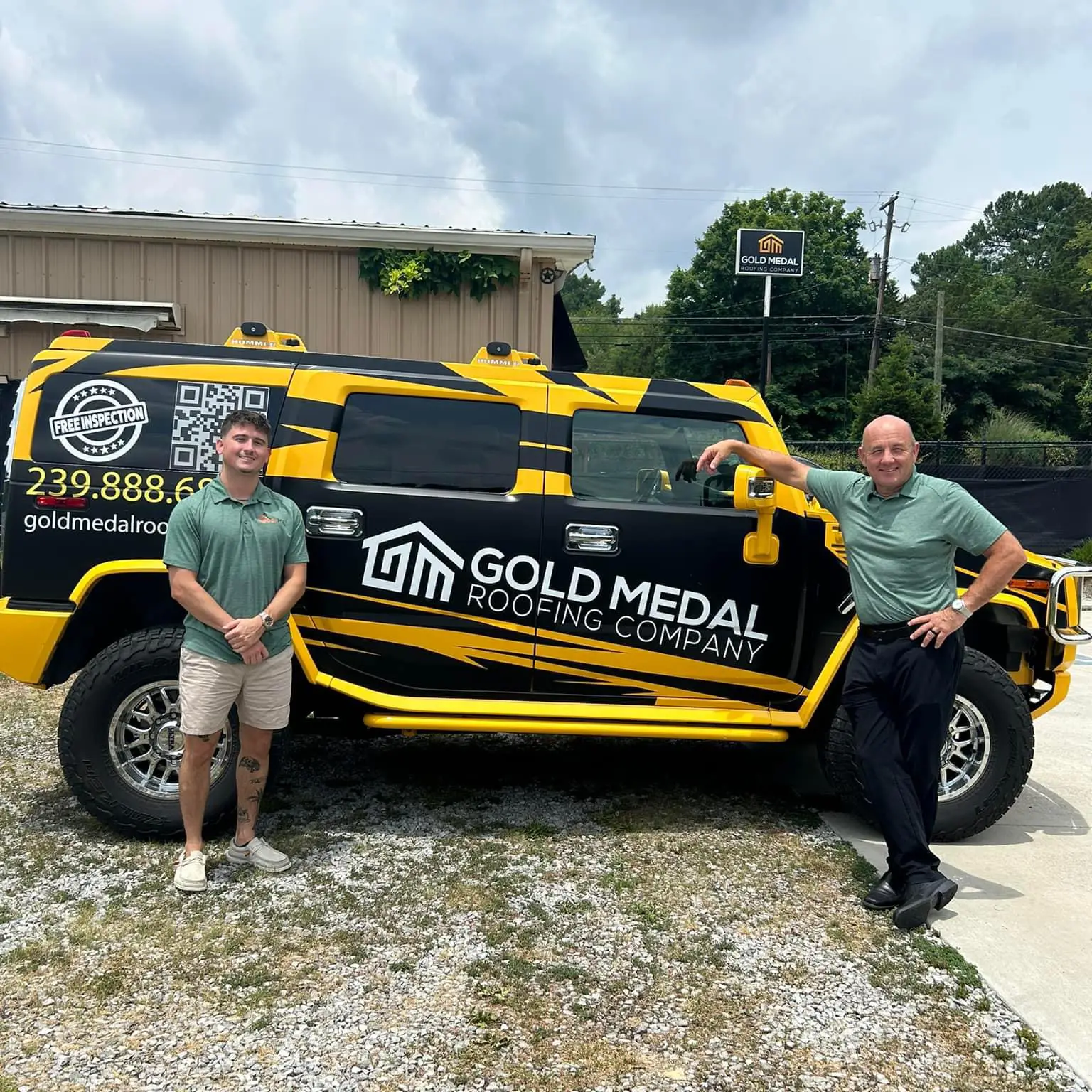 The image features two men standing in front of a black and yellow Hummer, showcasing the logo of Gold Medal Roofing Company. The man on the left, with short brown hair, wears a green polo shirt, khaki shorts, and white sneakers, while the man on the right, with a shaved head, sports a green polo shirt, black pants, and black shoes. The Hummer's side panel displays the company's logo, phone number, and website, accompanied by a QR code and the phrase "FREE INSPECTION." The vehicle is parked on a gravel lot, with a beige building and a black sign bearing the company's logo visible in the background. This image likely promotes the services of Gold Medal Roofing Company, a team of expert roofers based in Knoxville, Tennessee.
