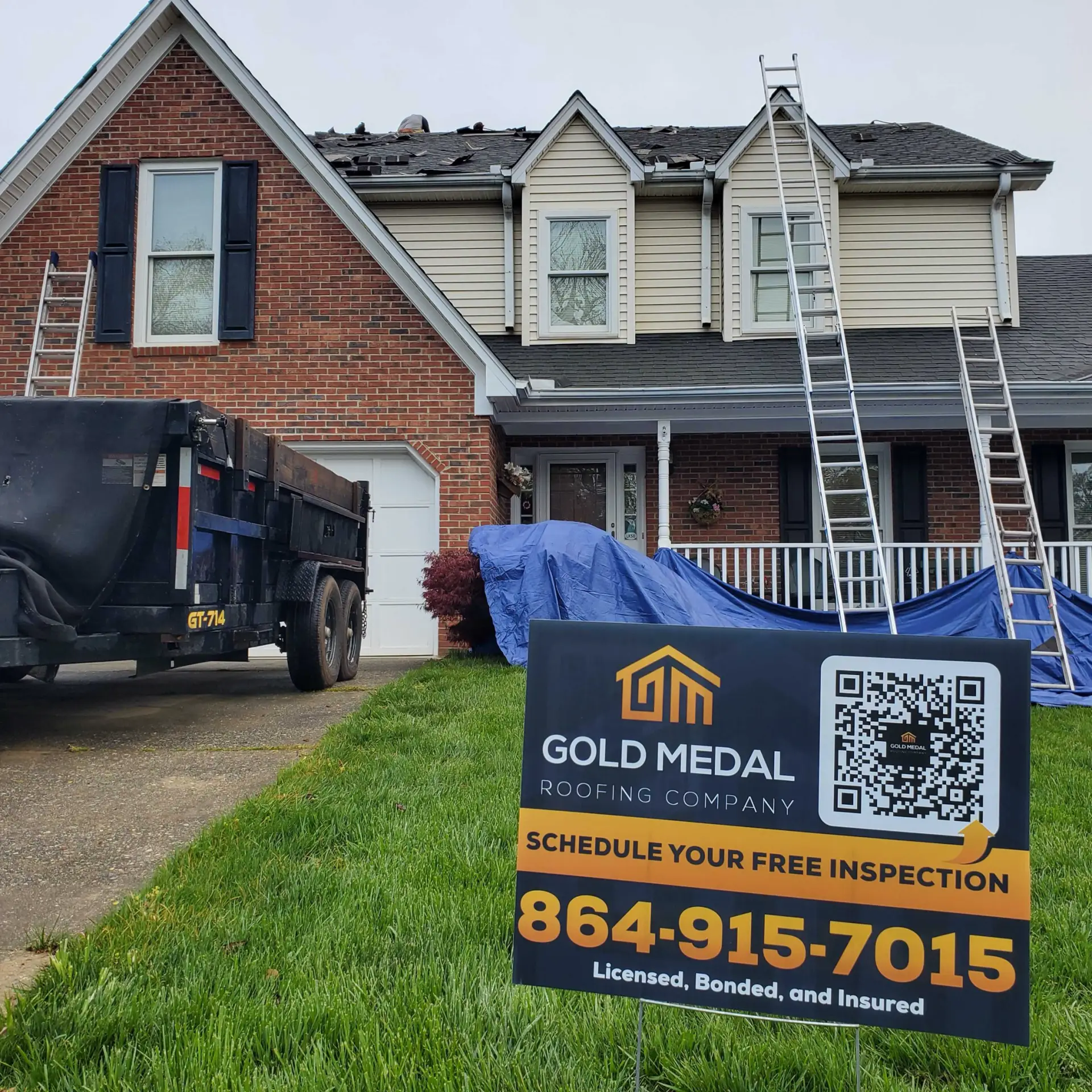 This image depicts a two-story house in Little Rock, Arkansas, with a prominent sign in the foreground advertising the services of Gold Medal Roofing Company. The sign, featuring a black background with white and gold text, includes a QR code and phone number, inviting viewers to "Schedule Your Free Inspection." In the background, a red brick house with beige siding is visible, with a tarp covering the front porch and ladders leaning against the roof, indicating that the roofers are actively working on the property. A black dump trailer is parked on the street to the left, adding to the scene's context.