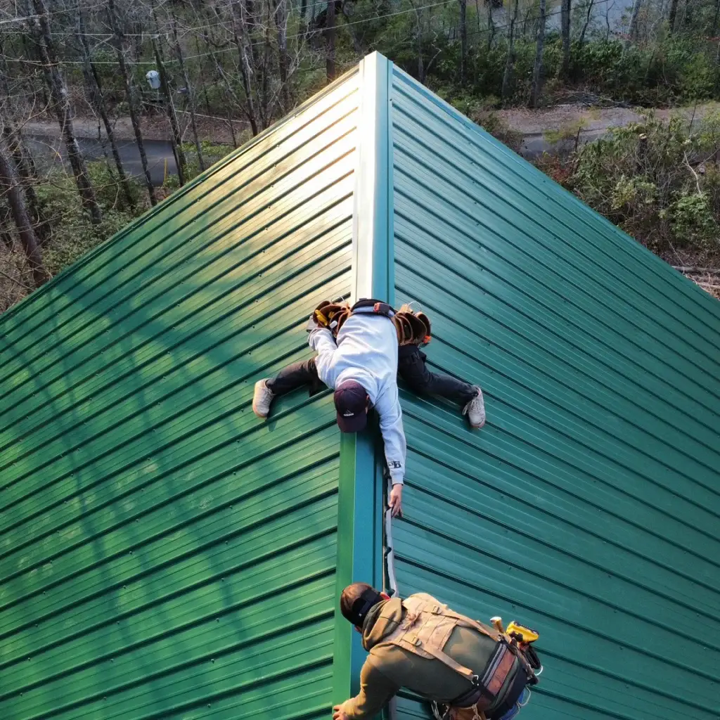 Two skilled workers from Gold Medal Roofing are captured in action, installing a sleek metal roofing system on a modern home. The image showcases the duo's expertise as they work in tandem, one kneeling on the roof's peak and the other standing on the edge, both clad in tool belts and safety harnesses. The metal roofing's green hue complements the surrounding wooded landscape, while the stone chimney and wooden deck add warmth to the scene. The image conveys a sense of professionalism and attention to detail, highlighting Gold Medal Roofing's commitment to quality craftsmanship.
