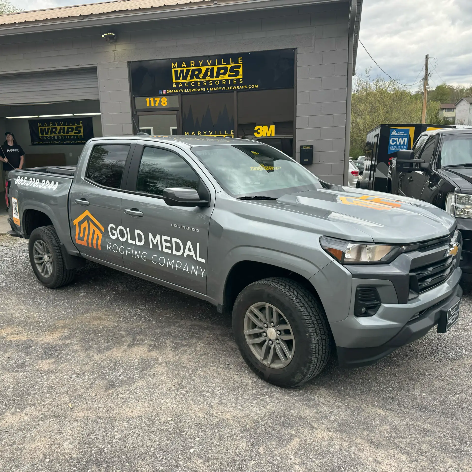 The image depicts a gray Chevrolet Colorado pickup truck parked in front of a building, prominently displaying the logo of Gold Medal Roofing Company on its side. The logo features a stylized orange roofline with the company name in white text, accompanied by a phone number. The truck is situated in a gravel parking lot, with a black pickup truck partially visible to its right. In the background, a gray brick building with a brown roof houses Maryville Wraps & Accessories, as indicated by a sign above the garage door. Two men are visible inside the building, one standing in the doorway and the other looking out the window. The scene is set against a backdrop of trees and houses, with a cloudy sky above. This image showcases the vehicle of a roofing company, likely used by roofers for their work.