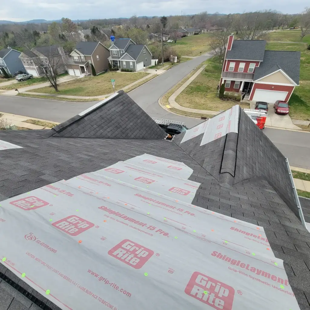 The image depicts a residential neighborhood with a prominent focus on a roof undergoing repair, contracted by Gold Medal Roofing. The roof, situated in the foreground, features gray shingles and is covered with white underlayment material bearing the "Grip Rite" logo in red. The surrounding neighborhood is characterized by a mix of two-story houses with varying exterior colors, including red, gray, and tan, and well-manicured lawns. A winding road traverses the neighborhood, with a few cars parked along the street. In the background, a grassy field stretches out, dotted with trees and a mountain range visible on the horizon under a cloudy sky.