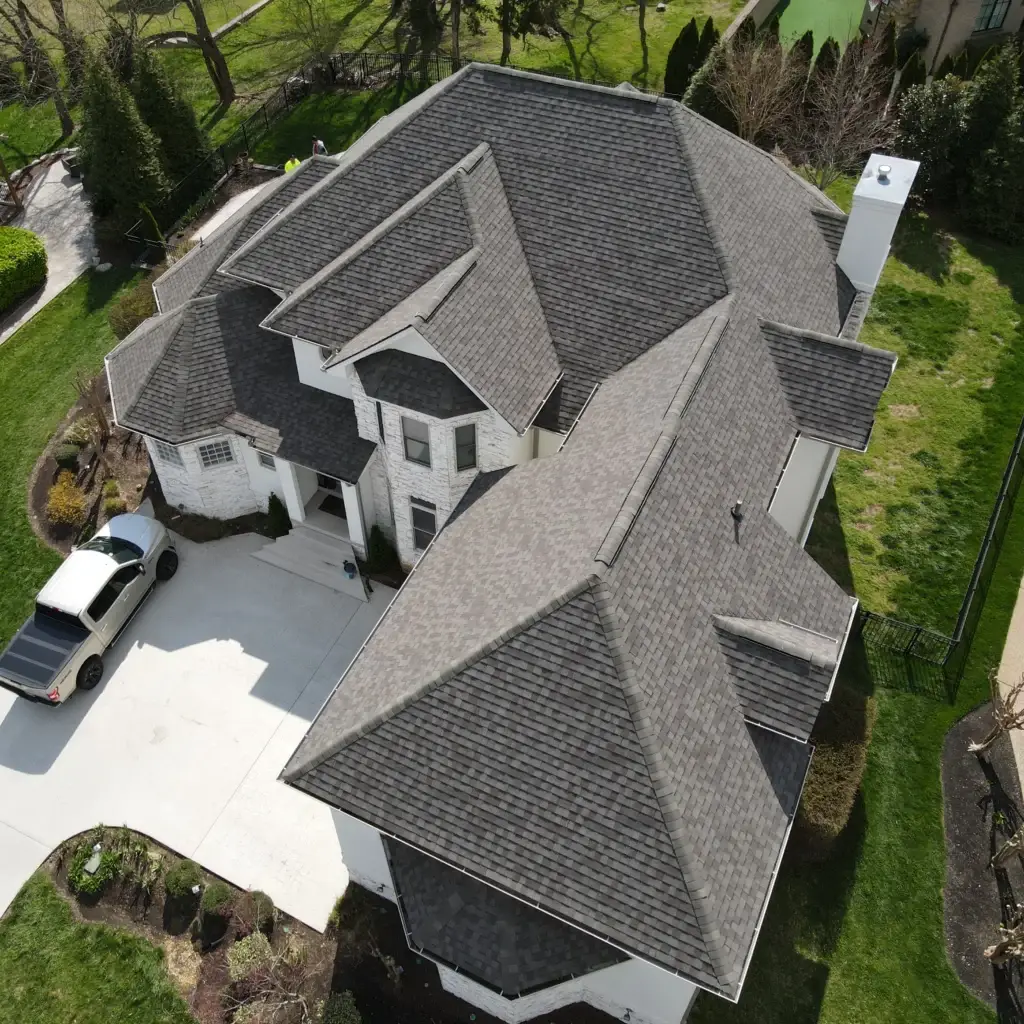 This aerial image showcases a two-story house with a gray roof, recently replaced by Gold Medal Roofing, as indicated by the presence of a white truck with a black bed cover in the driveway. The house features a white brick exterior, a chimney, and a large driveway leading to the front door. The surrounding landscape includes a well-manicured lawn, trees, and bushes, with neighboring houses and yards visible in the background. This image likely serves as a portfolio piece for Gold Medal Roofing, highlighting their expertise in roof replacement.
