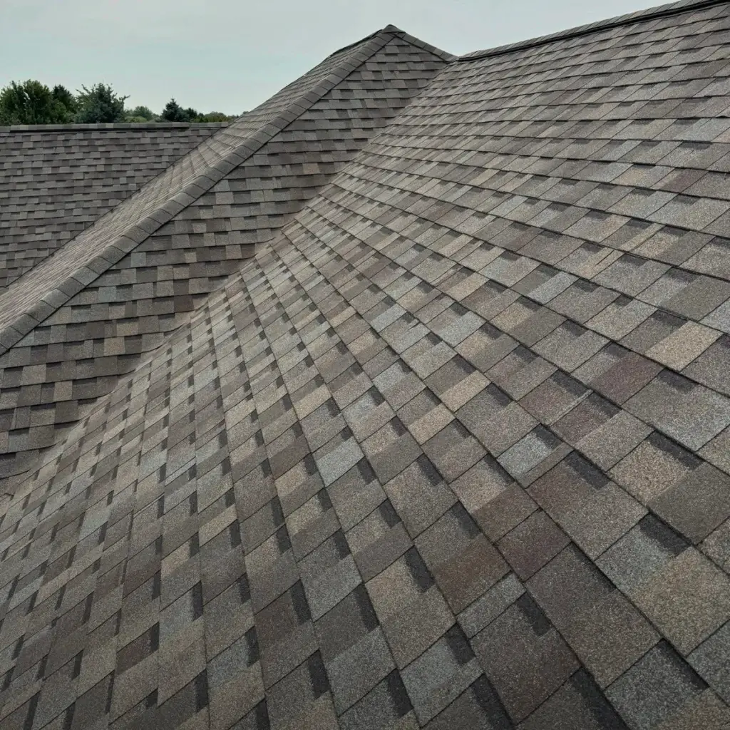 Newly installed shingle roofing showcases a harmonious blend of earthy tones, with a mix of brown, gray, and tan hues. The roof's steep pitch and multiple peaks create a visually appealing design, while the overcast sky provides a subtle backdrop. In the distance, lush green trees add a touch of natural beauty to the scene. Completed by Gold Medal Roofing, this image highlights the company's exceptional workmanship and attention to detail in shingle roofing installations.