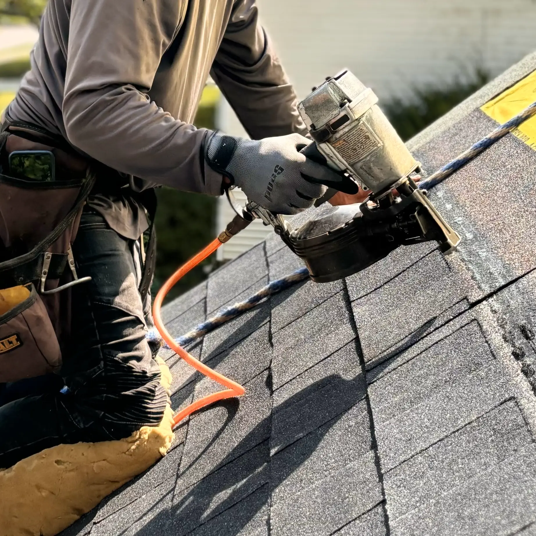 Roofer installs shingles with a nail gun on a roof being replaced by Gold Medal Roofing. He is kneeling on a cushion while working on the steep roof.